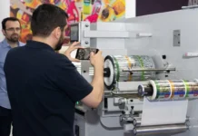 Gulf Print & Pack announces January 2027 dates Man photographing a large label-printing machine in an exhibition, rolls of labels feeding through the printer behind him.