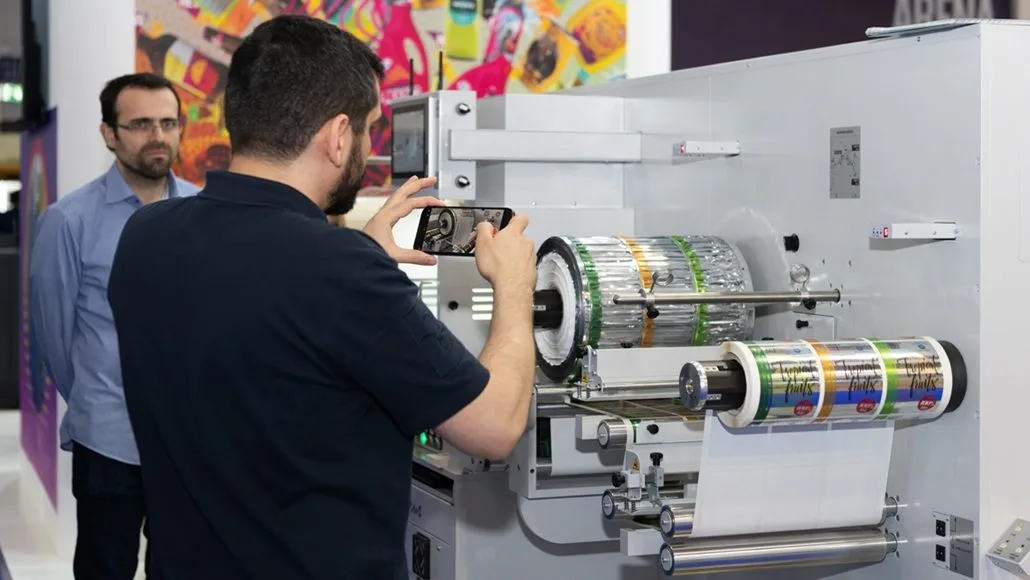Man photographing a large label-printing machine in an exhibition, rolls of labels feeding through the printer behind him.
