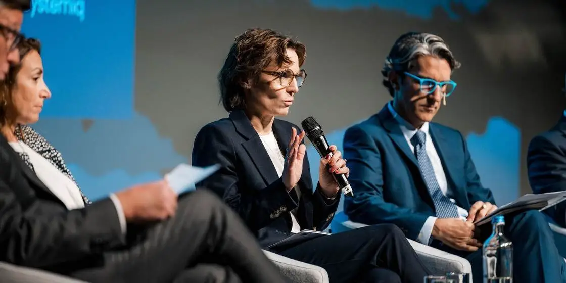 Sustainable-Packaging-Summit2026-El-1120x560-PWI Woman in a navy suit speaks into a handheld microphone during a panel discussion; colleagues listen attentively on either side in a formal setting with blue backdrop.