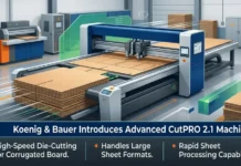 Koenig & Bauer Introduces Advanced CutPRO 2.1 Machine Factory floor with a large blue die-cutting machine cutting cardboard sheets; stacks of cardboard nearby. A banner reads: 'Koenig & Bauer Introduces Advanced CutPRO 2.1 Machine'.