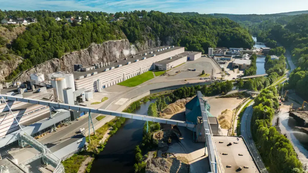 Aerial view of a large industrial quarry with long buildings, conveyors, and piles of material beside a winding river and green hills.