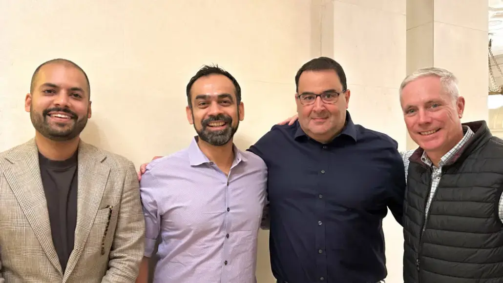 Four men stand side by side indoors, smiling at the camera with relaxed poses.