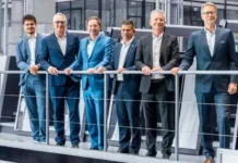 HEIDELBERG and pfenning Group Advance Pharma Packaging Six men in business suits lean on a railing in a modern manufacturing showroom or industrial space, posing for a group photo.