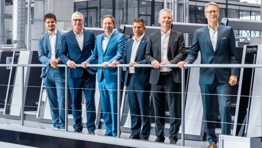 Six men in business suits lean on a railing in a modern manufacturing showroom or industrial space, posing for a group photo.